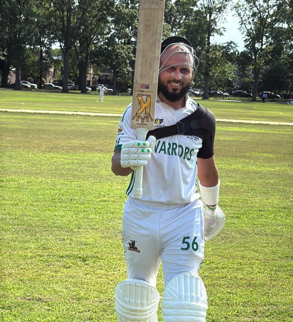 Cricketer holding a bat on a cricket field with trees and cars in the background