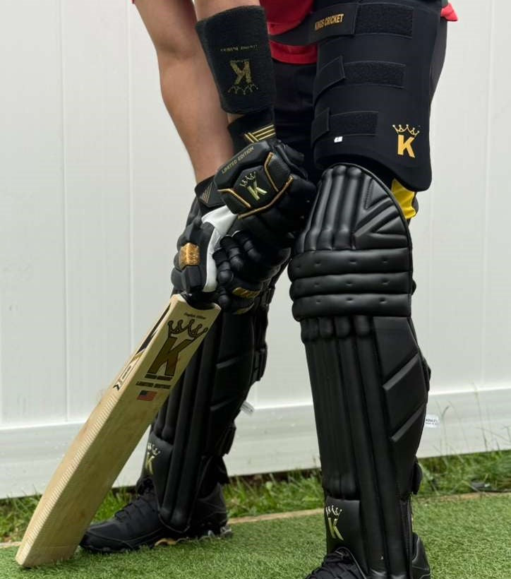 Person wearing black cricket gear with a bat, standing on grass against a white wall.